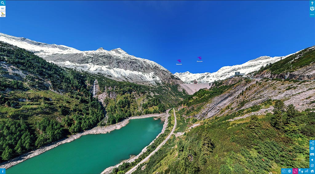 Das Bild zeigt eine 360° Panorama Aufnahme der Kölnbreinsperre. Links ein blauer Stausee, rechts grüne Wiesen und im Hintergrund die graue Staumauer- Darüber verschneite BErge und blauer Himmel.