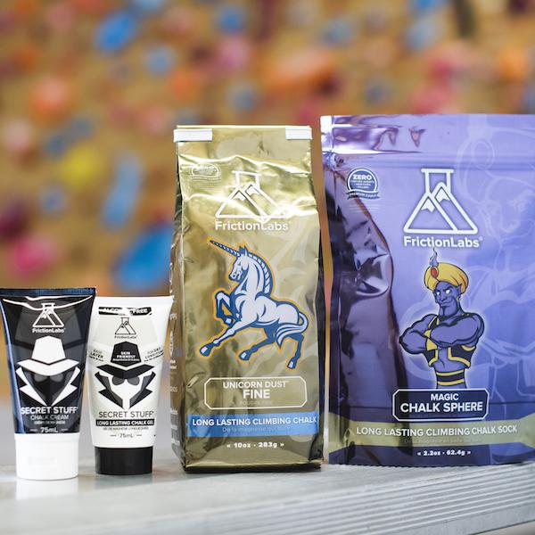 Das Bild zeigt vier Produkte des Chalk Herstellers Frictionabs. Von links nach rechts sind aufrecht stehend zu sehen: schwarze Tube Secret Stuff Liquid Chalk, weiße Tube Secret Stuff alkoholfrei Liquid Chalk, goldene Packung Unicorm Dust und violette Packung Magic Sphere Chalk Sock.