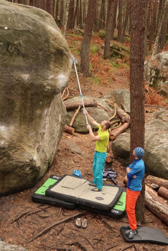 Das Bild zeigt ein Beispiel für den Boulderausrüstung Gegenstand Teleskop Putzstock. Ein Boulderer steht am Boden und putzt mit der Boulderbürste am Ende de Stocks einen Griff in ca. drei Metern Höhe.