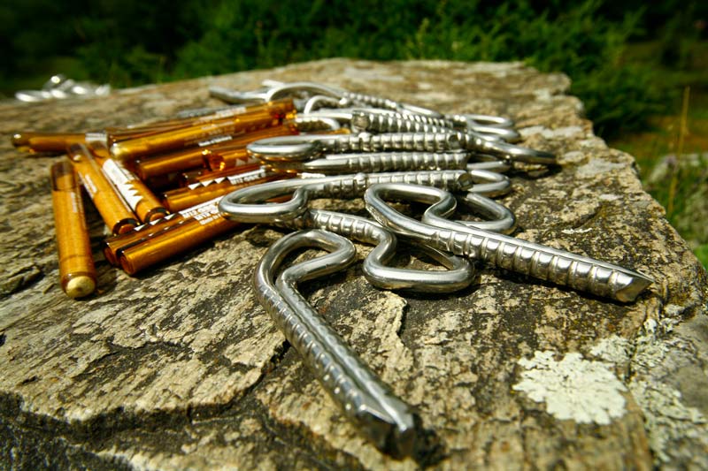 Das Bild zeigt einige silberne Klebehaken und braune Glasmörtelpatronen als Beipsiel für Einbohrartikel auf einer grauen Felsplatte.