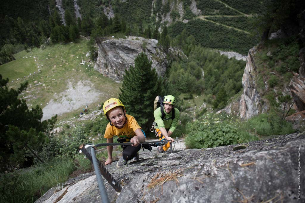 Das bild zeigt den Einsatz eines Klettersteigsets Kinder auf einem Klettersteig. An einer grauen Wand klettert ein Junge mit gelbem Helm voraus. Dahinter der Vater. im Hintergrund eine grüne Wiese bzw. Wald.
