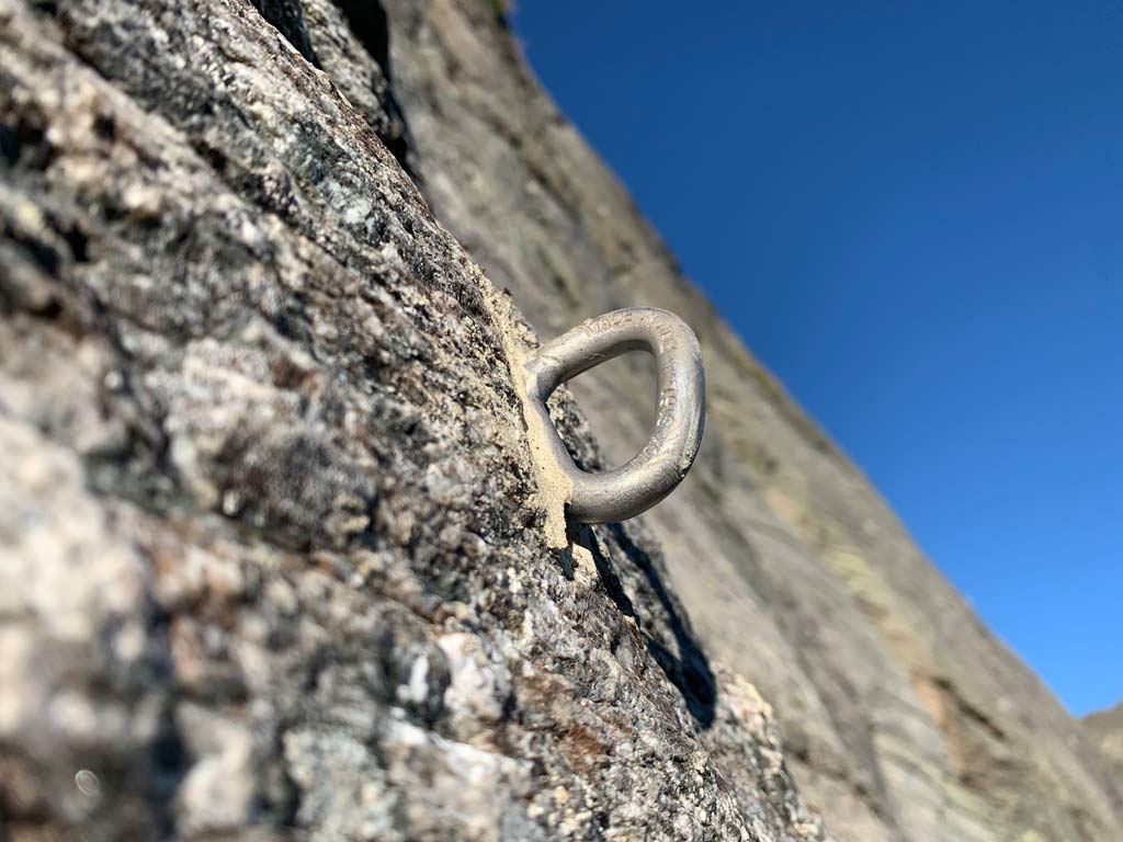 Das Bild zeigt einen perfekt gesetzten Klebehaken bei einer Klettergarten Sanierung. Er steht aus grauem Urgestein hervor. Das Bild ist im Vordergrund bewusst unscharf, damit der gestochen scharfe Haken ins Zentrum des Blickes rückt. im Hintergrund blauer Himmel.