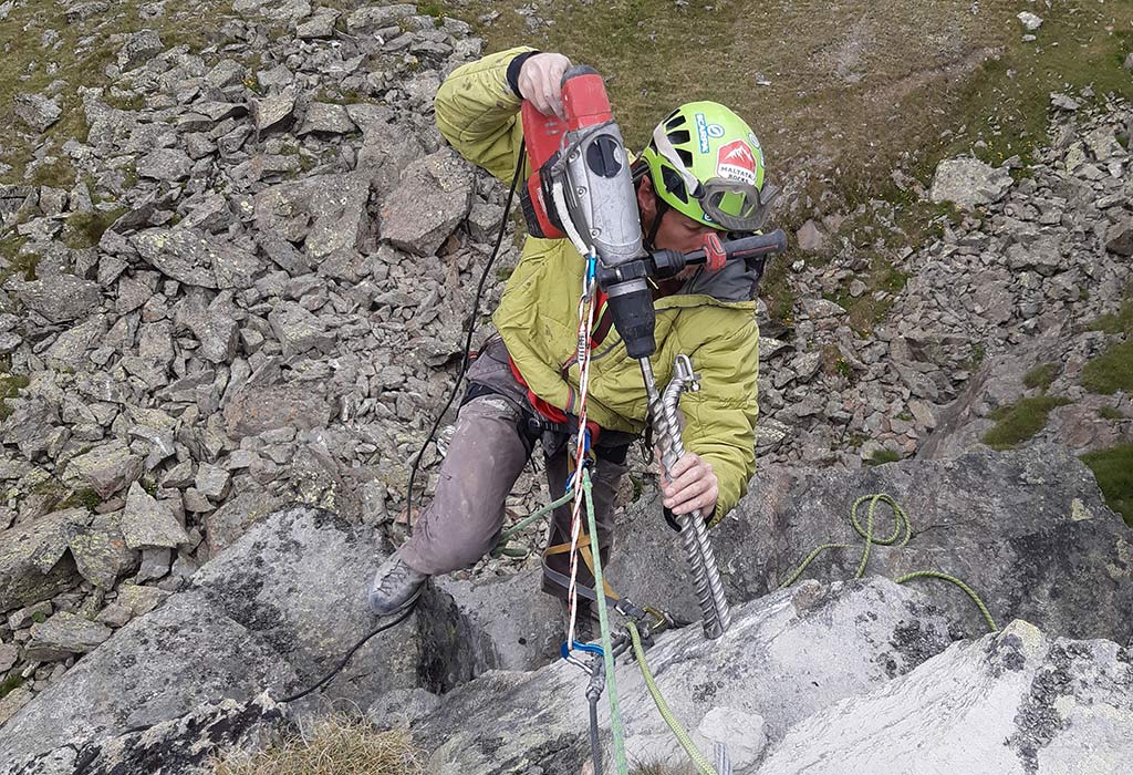 Das Bild zeigt einen arbeiter der als Beispiel für eine wichtige Kletter Infrastruktur einen Klettersteig errichtet. Er hängt im Seil und hält einen großen Bohrhammer mit einem Klettersteiganker an den Felsen.