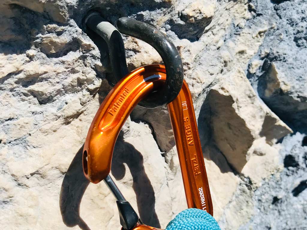 Das Bild zeigt einen Lusterhaken in einer Felswand mit orangem, eingehängten Karabiner. Das Set up stellt eine potentiell tödliche, komplett inadäquate Absicherung für Kletterrouten dar!