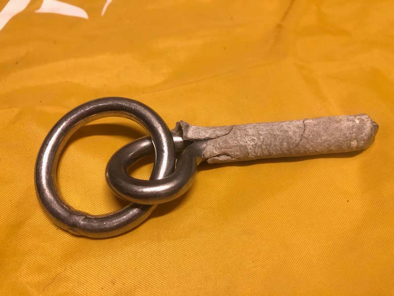 Das Bild zeigt einen Klebehaken mit Ring aufeiner gelben Plane. Um den Hakenschaft ein pfropfen aus Mörtel.